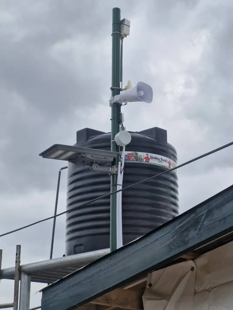 A pole carrying a camera, horn speaker, solar light and antenna beside a water tank.