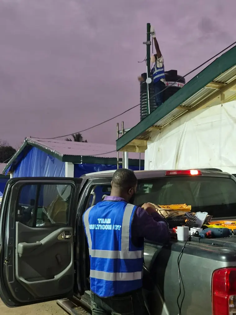 A MulticomIoT technician working from a vehicle while a colleague installs equipment on a roof at dusk.